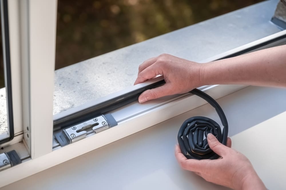 A woman sticks a dark rubber sealing tape on a window indoors to save energy